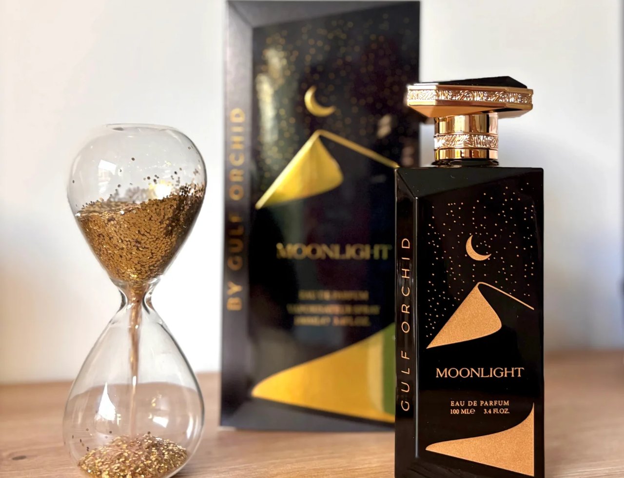 Gulf Orchid Moonlight Eau De Parfum 100ml by Gulf Orchid — Botol minyak wangi hitam berkilau dengan penutup emas, di sebelah jam pasir berisi glitter emas dan kotak minyak wangi.