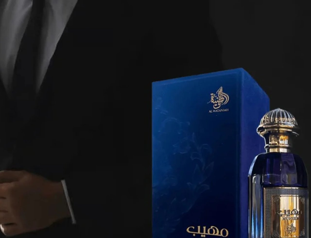 Al Wataniah Muheeb Perfume Man by Al Wataniah 100ml — Kotak biru gelap dengan botol minyak wangi berpenutup emas berkilat, latar belakang gelap dengan sut lelaki