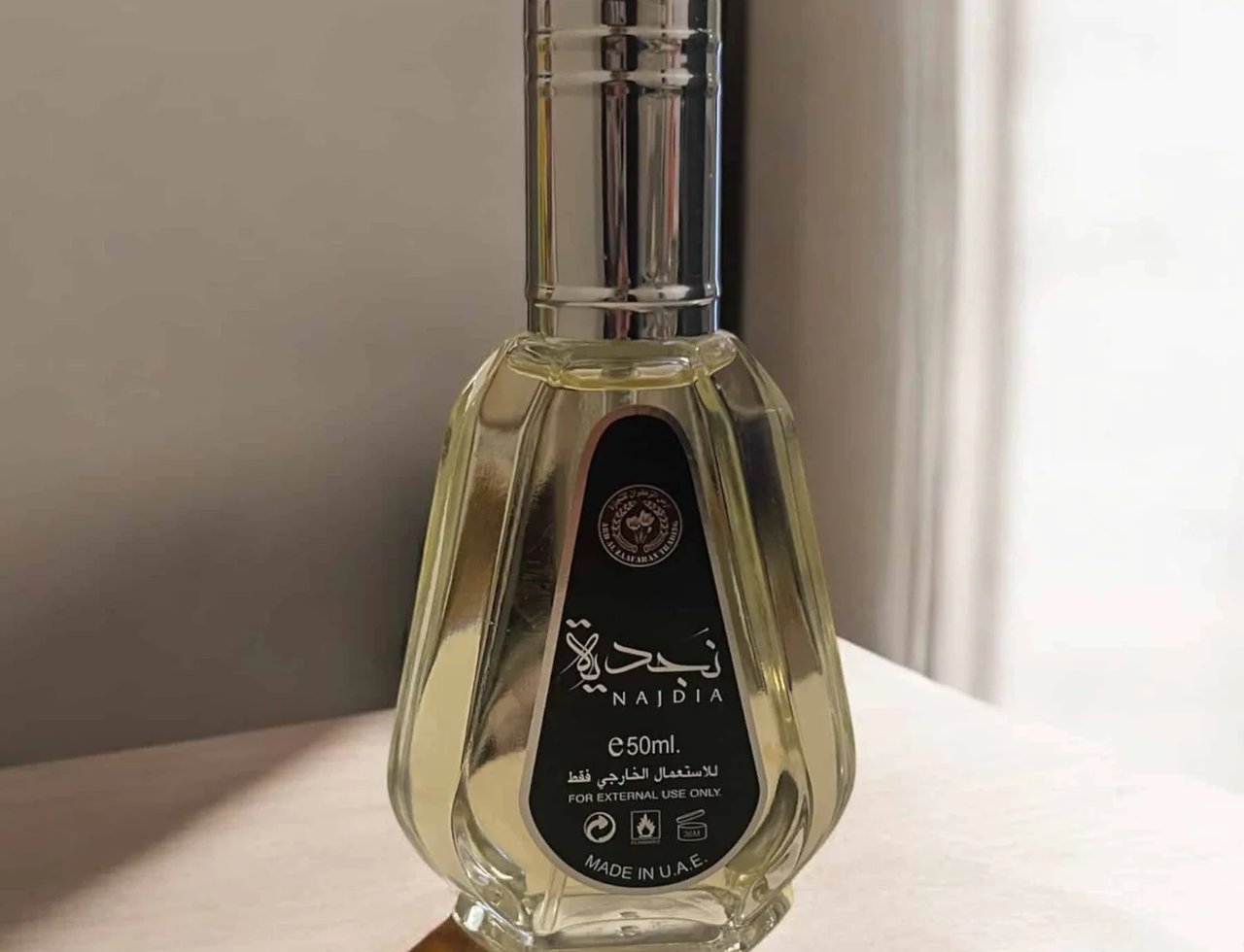 Ard Al Zaafaran Najdia by Ard Al Zaafaran 50ml EDP — Botol minyak wangi kaca jernih dengan penutup perak, berlabel hitam bertulis Arab dan Inggeris, diletakkan di atas permukaan cerah dengan bayang.