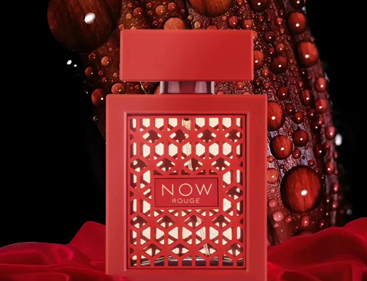 Rave Now Rouge Perfume EDP 100ml by RAVE — A red perfume bottle with a geometric lattice design sits on red satin fabric, with a dark background featuring water droplets on a textured surface. | Rave Now Rouge Perfume EDP 100ml by RAVE — Botol minyak wangi merah dengan reka bentuk krat geometri diletakkan di atas