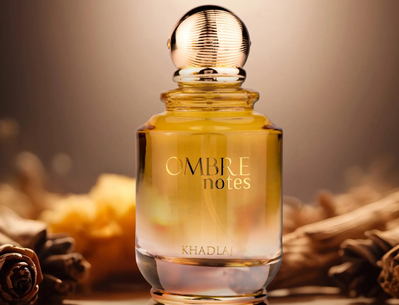 Khadlaj Ombre Notes by Khadlaj 100ml EDP Unisex — Botol minyak wangi kaca jernih berisi cecair keemasan dan penutup emas berjalur, berlatar belakang coklat gelap kabur dengan elemen semula jadi seperti kayu dan rempah.