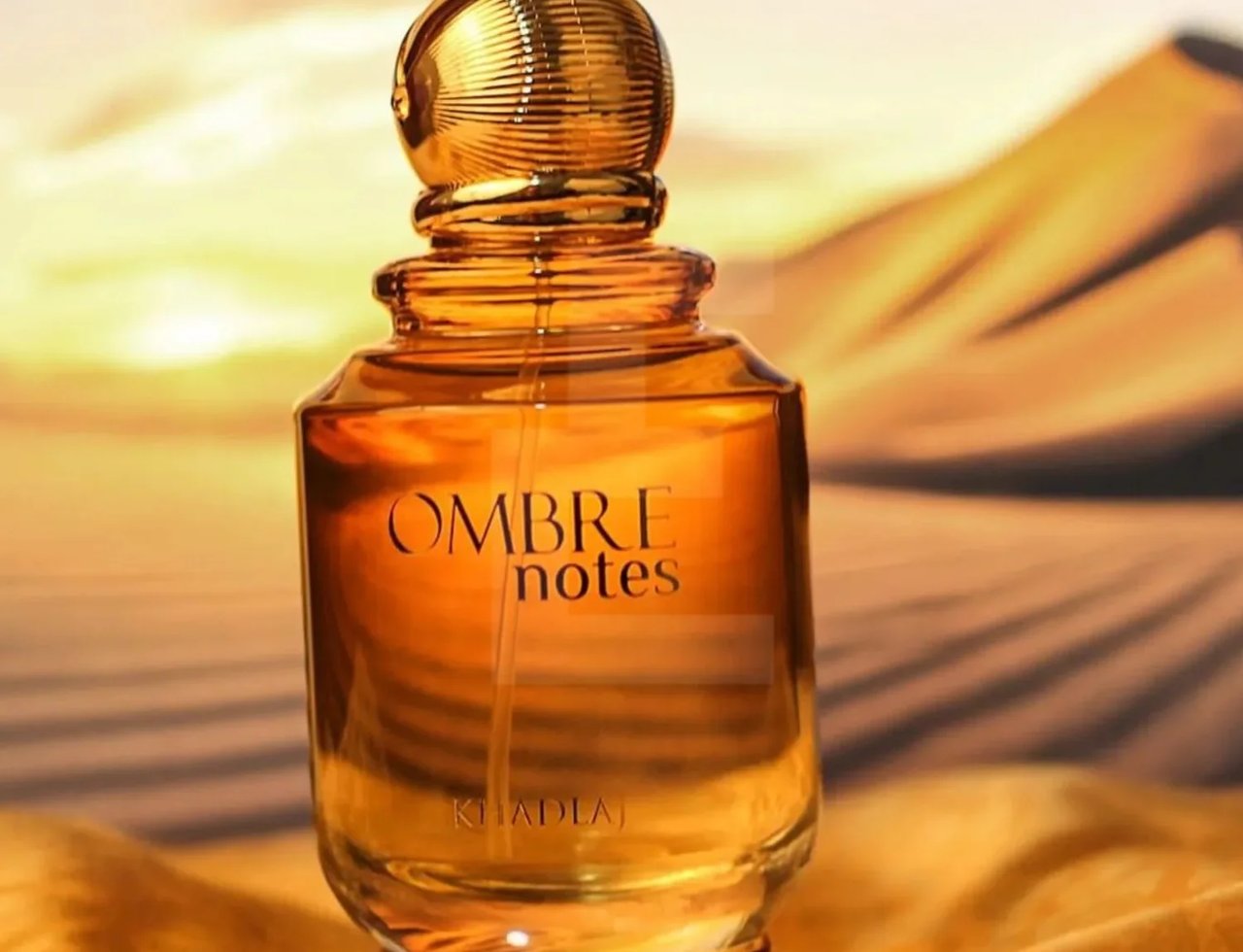 Khadlaj Ombre Notes by Khadlaj 100ml EDP Unisex — Amber glass perfume bottle with a textured gold cap, set against a desert landscape at sunset.