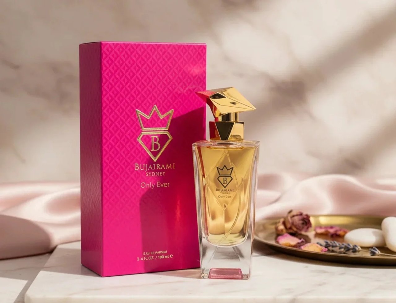 Bujairami Only Ever by Bujairami 100ml EDP — Botol minyak wangi kaca bertutup emas di sebelah kotak merah jambu terang bertekstur, diletakkan di atas permukaan marmar bersama fabrik sutera dan bunga kering hiasan.