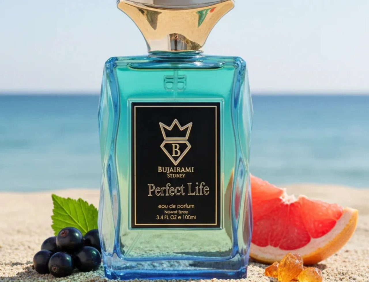 Bujairami Perfect Life Eau de Parfum 100ml — Blue glass perfume bottle with gold cap on a sandy beach with ocean background, featuring grapefruit slice and blackcurrants.