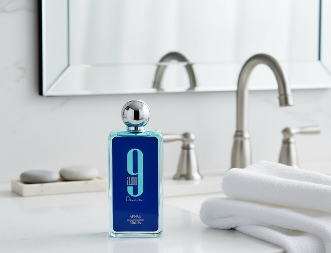 Afnan 9AM Dive by Afnan EDP 100 ml For Men — Botol wangian biru terang dengan penutup bulat berkilat, diletakkan di atas meja singki bilik air dengan cermin dan pili air di latar belakang, serta tuala putih dilipat di sebelah kanan.