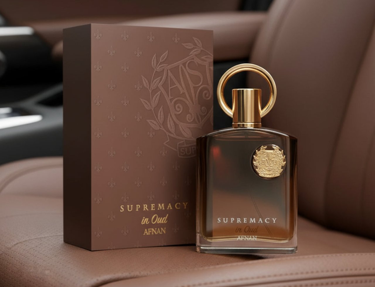Afnan Supremacy In Oud by Afnan Perfumes – Kotak coklat bertekstur dengan corak bunga fleur-de-lis dan logo berukir, bersebelahan dengan botol kaca coklat gelap dengan penutup dan cincin emas, diletakkan di atas tempat duduk kereta kulit coklat.
