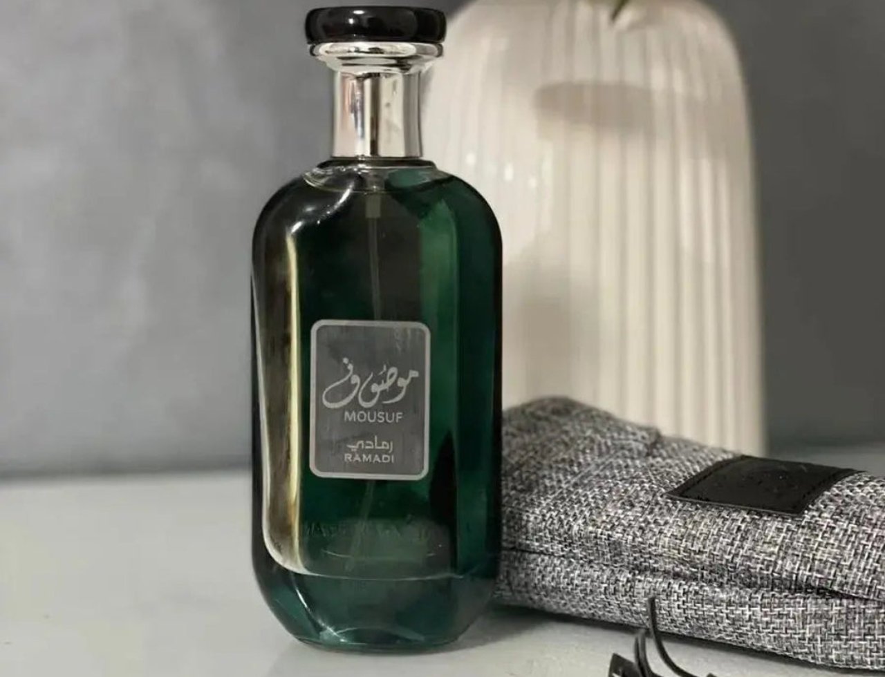 Ard Al Zaafaran Mousuf Ramadi by Ard Al Zaafaran 100 ml for Men — Botol minyak wangi hijau gelap dengan label perak, penutup hitam, di atas meja putih berlatar belakang pasu bunga putih dan kain kelabu