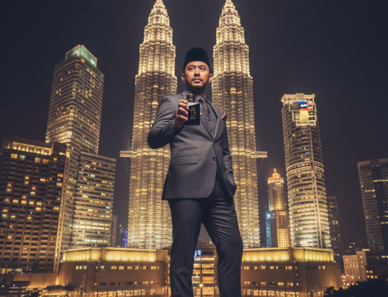 Ard Al Zaafaran Oud 24 Hours Ard Al Zaafaran 100ml EDP — Lelaki berbaju Melayu lengkap dengan songkok memegang botol wangian di hadapan bangunan berkembar Petronas pada waktu malam.