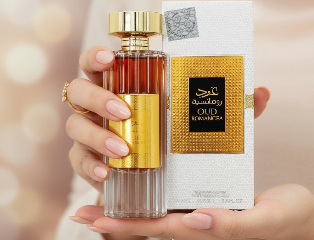 Ard Al Zaafaran Oud Romancea by Ard Al Zaafaran 100ml for Women — Tangan wanita memegang botol minyak wangi kaca lutsinar dengan cecair perang, penutup dan jalur emas berkilat, bersama kotak putih bertekstur dengan perincian emas.