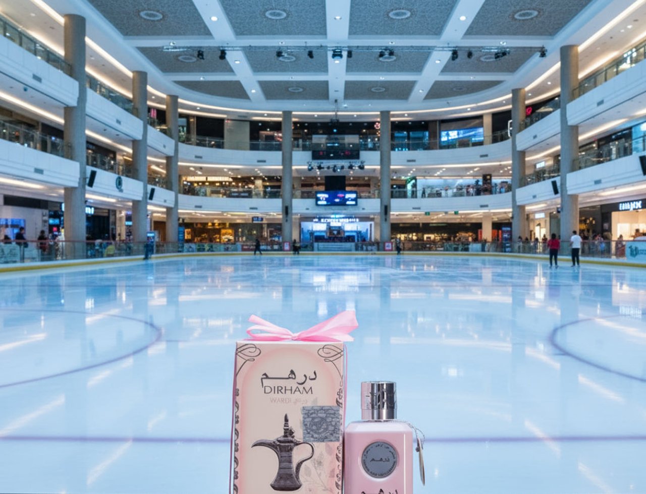 Ard Al Zaafaran Dirham Wardi — Pink perfume bottle and box on a dark surface in front of an indoor ice rink.