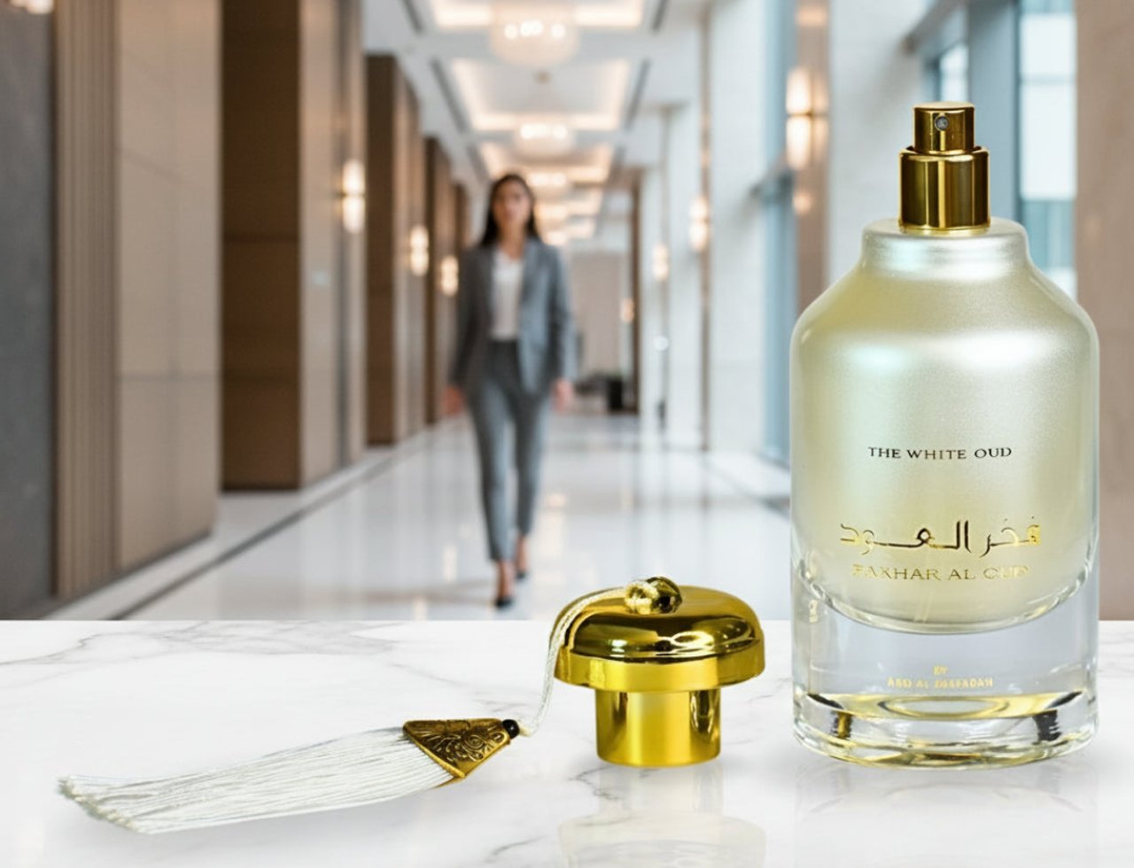 Ard Al Zaafaran Fakhar Al Oud “The White Oud” — Perfume bottle and gold cap with tassel on marble, blurred woman in suit