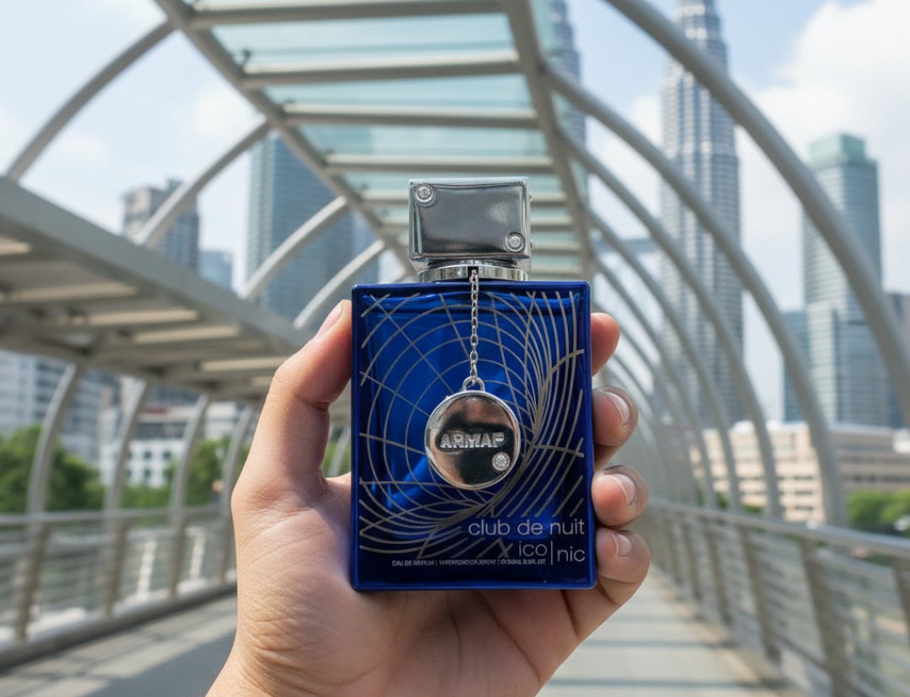 Armaf Club De Nuit Iconic 105ml EDP Men — Tangan memegang botol wangian biru gelap dengan penutup perak dan loket bulat, latar belakang jambatan moden dan bangunan pencakar langit