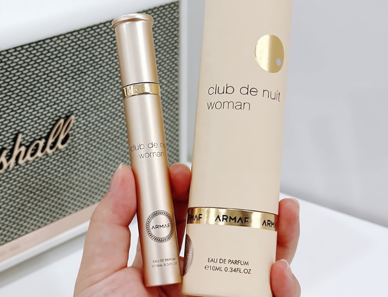 Armaf Club De Nuit Woman 10ml EDP — Tangan memegang botol semburan emas dan kotak panjang berwarna krim, latar belakang pembesar suara Marshall