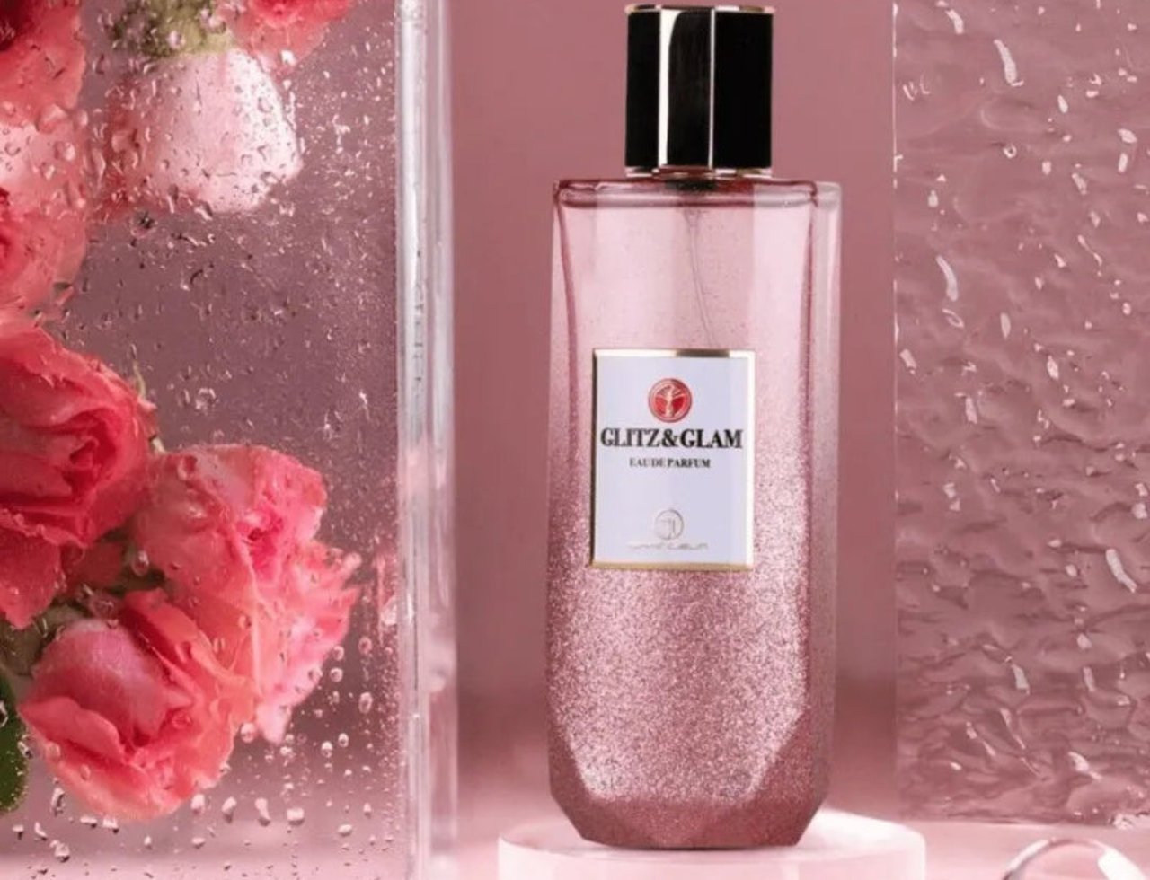 Grandeur Glitz Glam by Grandeur Elite 100 ml untuk Wanita — Botol minyak wangi pink berkilauan tinggi dengan penutup hitam beraksen emas, dipamerkan di atas tapak akrilik jernih. Mawar merah jambu bertitisan air kelihatan dalam bekas kaca di sebelah kiri.