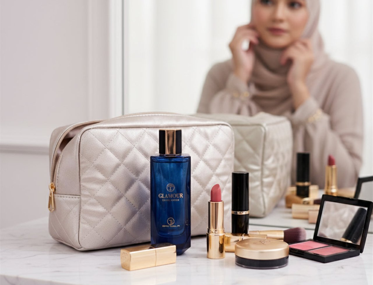 Grandeur Glamour Woman by Grandeur Elite 100ml — A blue perfume bottle with gold accents sits on a marble surface next to a quilted gold makeup bag, lipsticks, and a blush palette, with a woman in a hijab reflected in the background mirror.