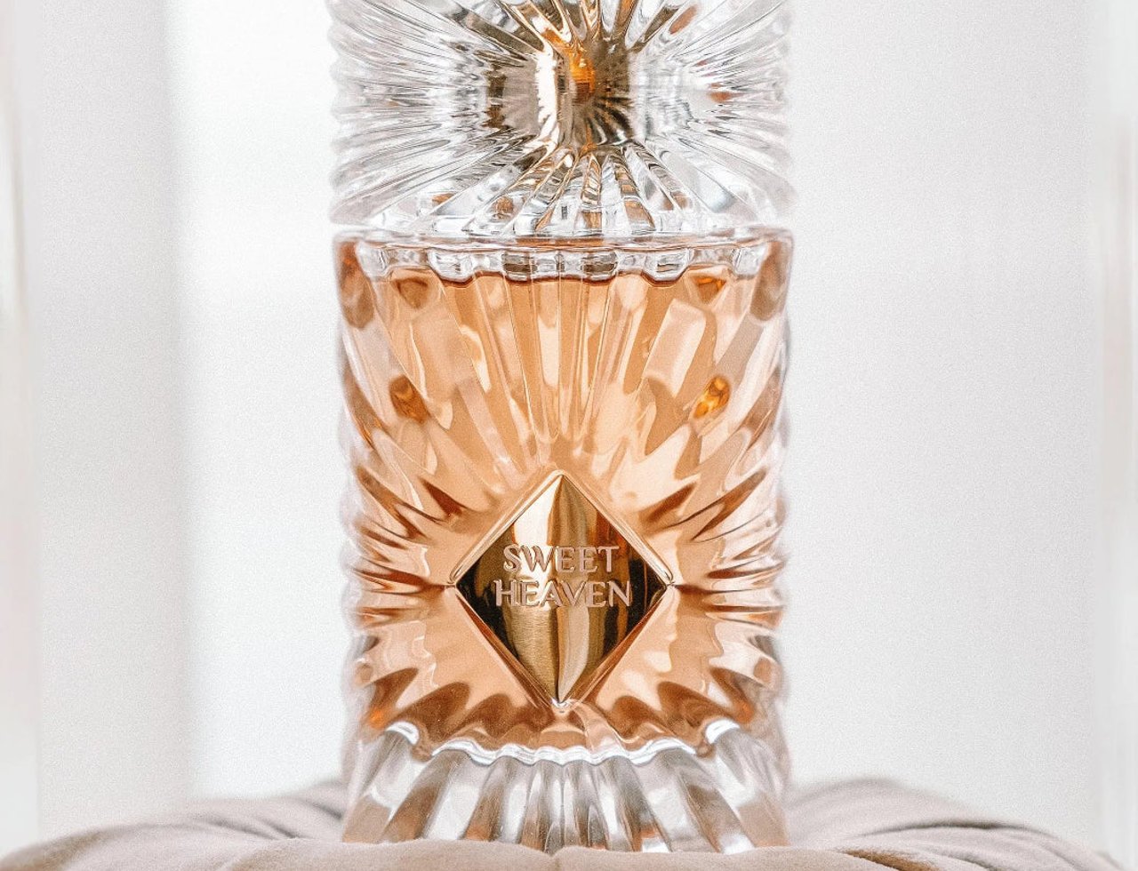 Gulf Orchid Sweet Heaven by Gulf Orchid 100ml EDP Unisex — Close-up of a clear, faceted glass perfume bottle with rose-gold liquid, featuring a diamond-shaped gold label with 'SWEET HEAVEN' text, resting on a soft, beige cushion.