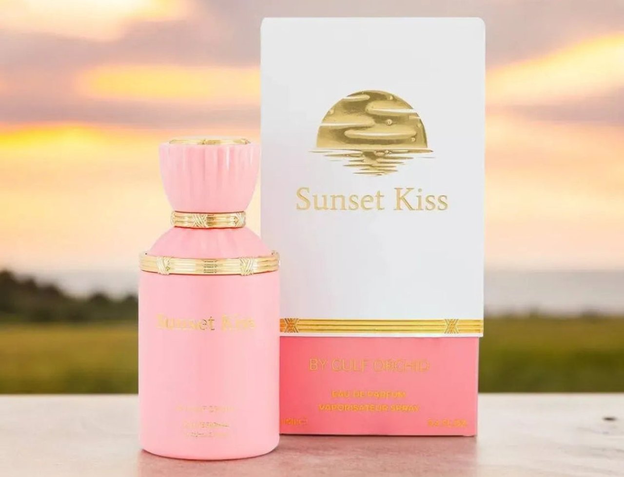 Gulf Orchid Sunset Kiss 100ml EDP Woman — Botol minyak wangi pink dengan perincian emas dan penutup berlekuk, bersebelahan kotak putih dan pinknya dengan logo matahari emas, berlatar langit senja.