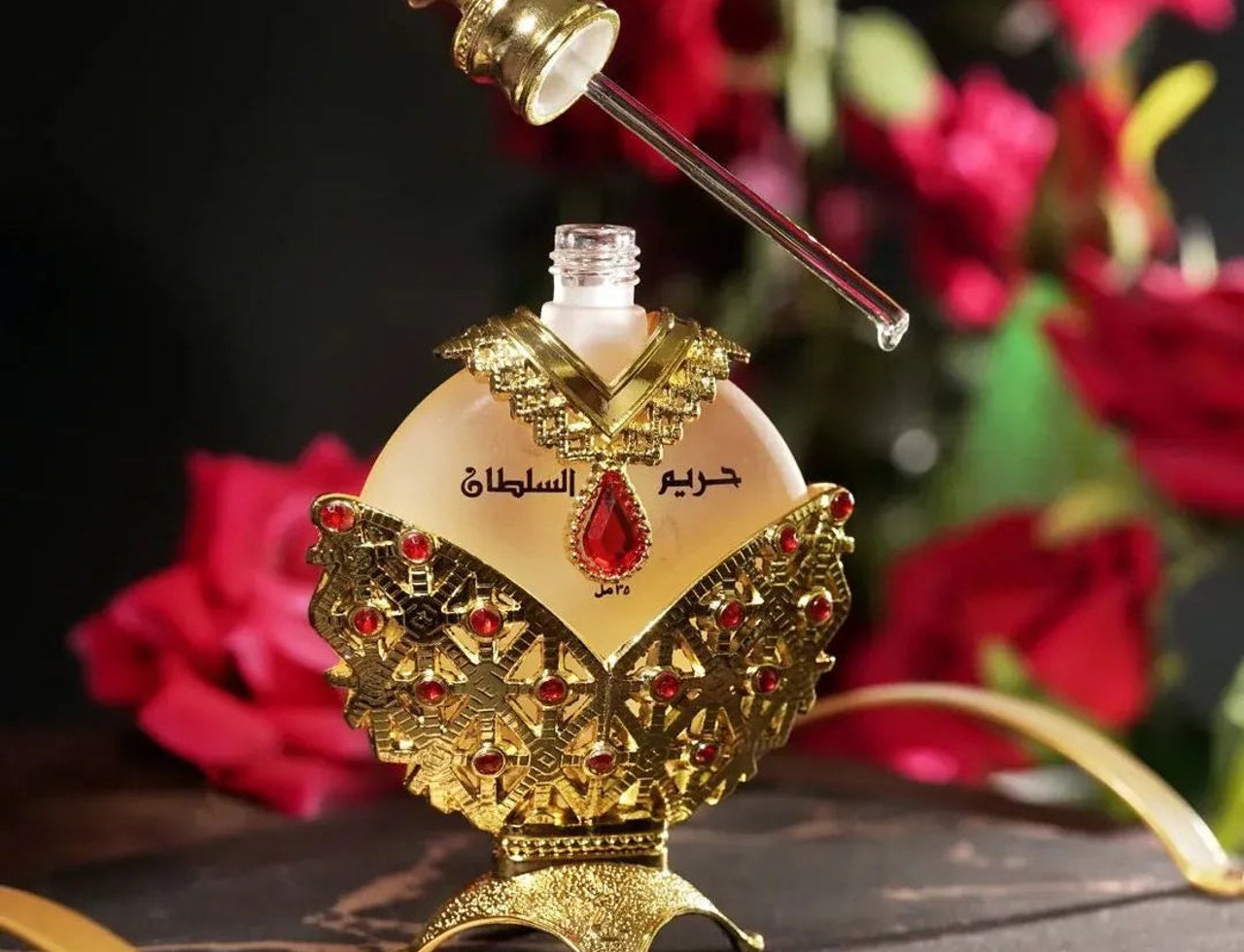 Khadlaj Hareem Al Sultan Gold by Khadlaj 35ml — Botol minyak wangi emas berhias dengan perincian permata merah, menampilkan penitis dengan setitik cecair, berlatar belakang merah jambu merah yang kabur.