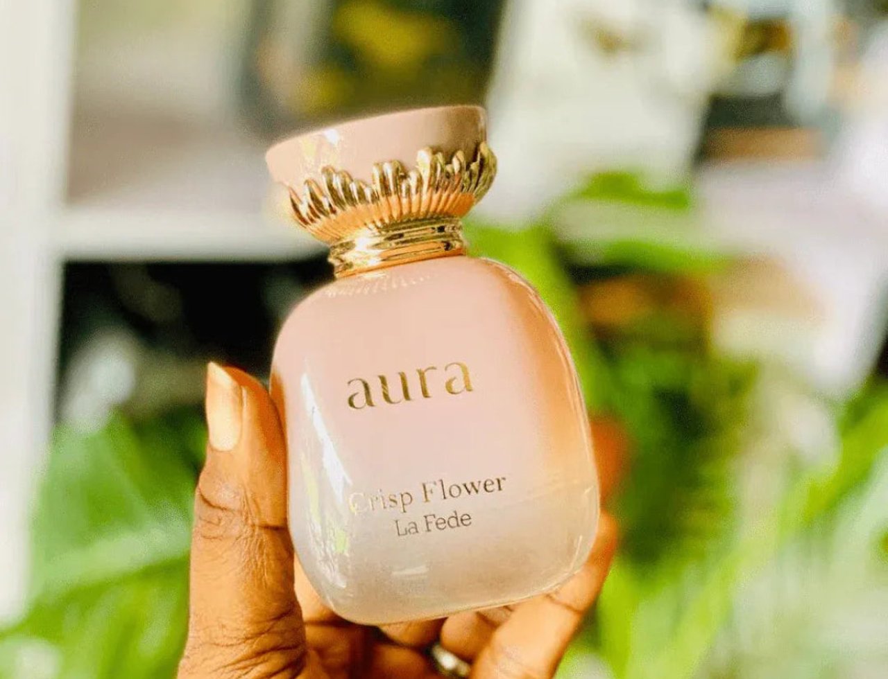 Khadlaj La Fede Elegant Crisp Flower 100ml EDP — Tangan memegang botol wangian berwarna merah jambu pucat dengan penutup emas berhias, berlatar belakang kehijauan daun yang kabur.