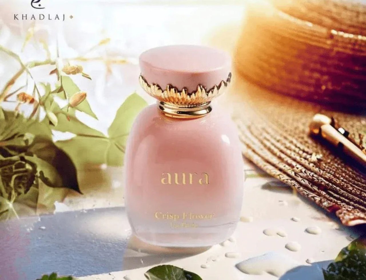 Khadlaj La Fede Elegant Crisp Flower 100ml EDP — Pink perfume bottle with gold ornate cap, surrounded by green leaves and water droplets, with a golden woven accessory in the background.