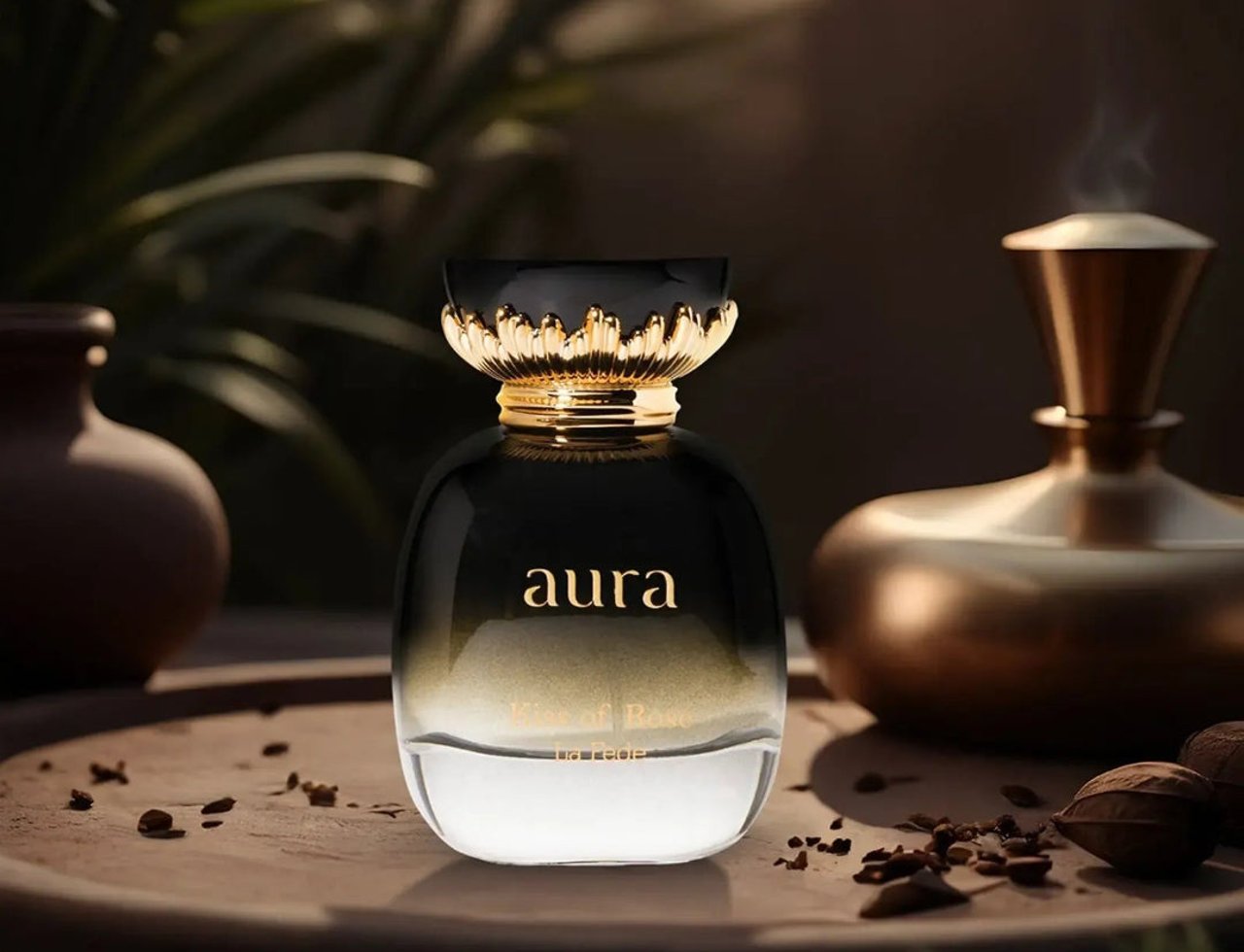 Khadlaj La Fede Elegant Kiss of Rose 100ml EDP — Botol minyak wangi gradien hitam emas dengan tulisan 'aura' dan 'Kiss of Rose', diletakkan di atas dulang kayu bersama kacang dan rempah bertabur, dengan pasu kabur dan penyebar emas di latar belakang.