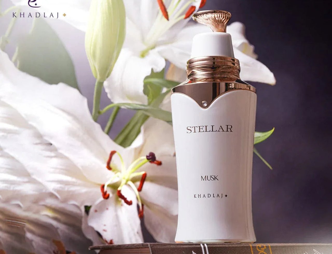 Khadlaj Stellar Musk by Khadlaj 100ml EDP — Botol putih dengan aksen emas dan penutup, dikelilingi bunga lily putih dan diletakkan di atas buku antik.