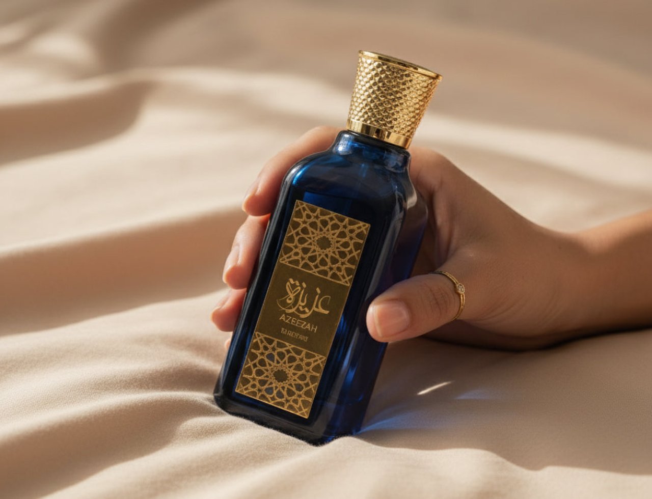 Lattafa Azeezah by Lattafa 100ml for Women — Tangan memegang botol kaca biru gelap dengan penutup emas bertekstur, label emas dengan corak geometri dan tulisan Arab, latar belakang kain berlipat warna krim.