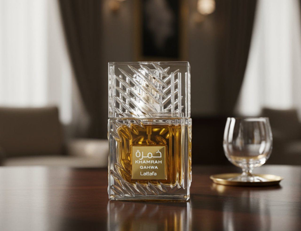 Lattafa Khamrah Qahwa by Lattafa 100ml EDP Unisex — Botol minyak wangi kaca jernih berisi cecair keemasan, bertudung kaca bertekstur, terletak di atas meja kayu berkilat bersebelahan gelas kecil di atas alas emas. Latar belakang adalah dalaman yang kabur lembut dengan langsir dan lampu dinding.