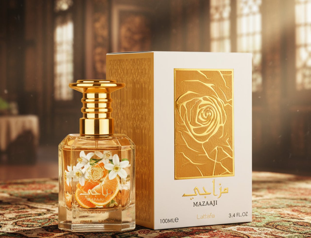 Lattafa Mazaaji by Lattafa 100ml EDP for Women — Clear glass perfume bottle with gold cap, orange slices, and white flowers, next to a gold and white box with a rose design, on a patterned rug.