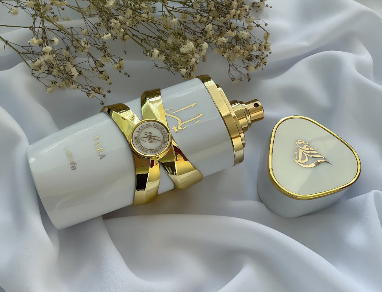 Lattafa Yara Moi by Lattafa 100ml EDP Women — White perfume bottle with gold accents and Arabic script, lying on white fabric next to its triangular gold-rimmed cap and dried flowers.