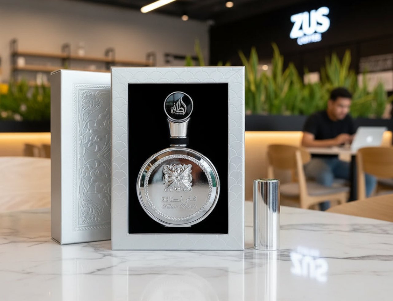 Lattafa Fakhar Platin — Silver perfume bottle and box displayed on a marble table with a blurred coffee shop background.