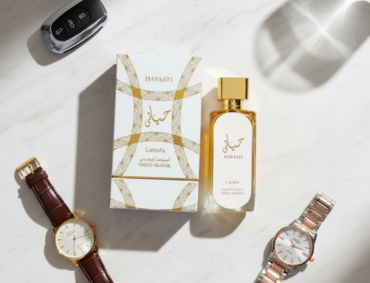 Lattafa Hayaati Gold Elixir by Lattafa 100ml Unisex — White and gold patterned box and bottle of Hayaati Gold Elixir perfume, with a car key and two watches on a marble surface.