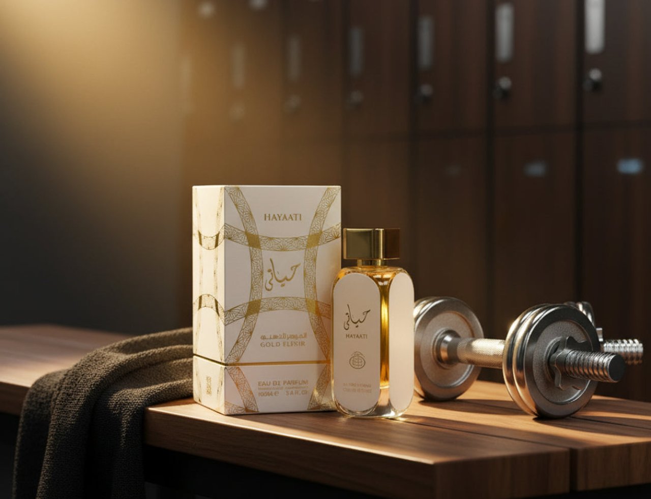 Lattafa Hayaati Gold Elixir by Lattafa 100ml Unisex — Kotak dan botol minyak wangi putih beraksen emas diletakkan di atas bangku kayu bersebelahan tuala gelap dan dumbbell, dengan loker gim di latar belakang.