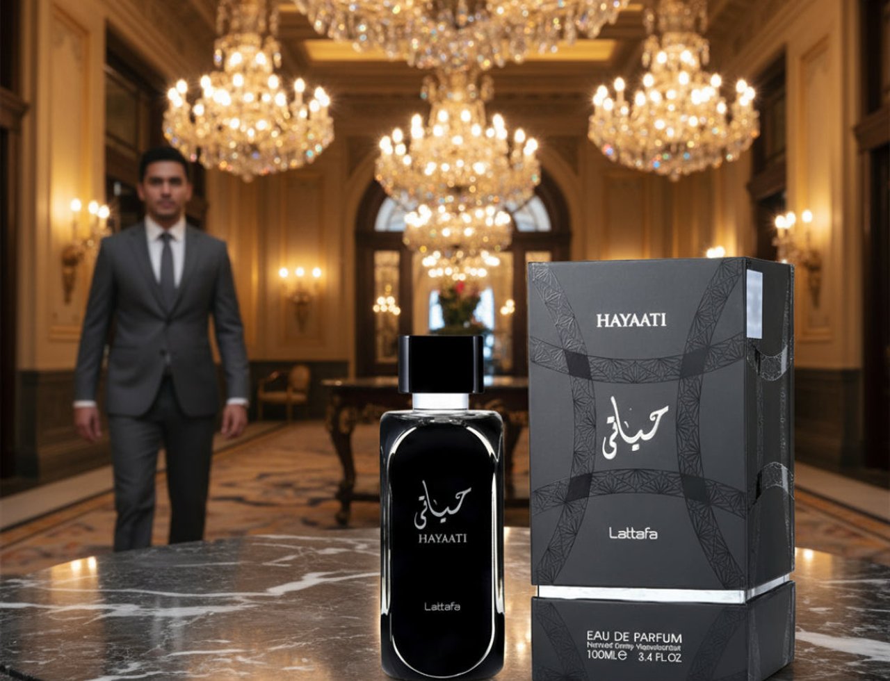 Lattafa Hayaati Men by Lattafa Perfumes 100ml EDP — Seorang lelaki berbaju kelabu lalu di hadapan botol minyak wangi Hayaati dan kotaknya, diletakkan di atas meja marmar di dewan mewah dengan candelier.