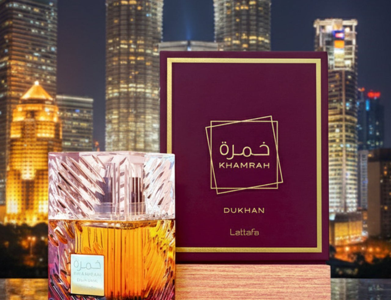 Lattafa Khamrah Dukhan by Lattafa 100ml EDP Unisex — Botol minyak wangi kaca jernih berisi cecair perang duduk di atas permukaan marmar, bersebelahan kotak merah marun dan emasnya, dengan latar belakang pemandangan bandar pada waktu malam.