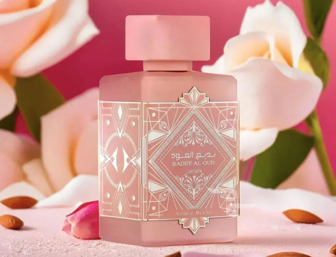 Lattafa Badee Al Oud Noble Blush — Pink glass perfume bottle with ornate geometric label, surrounded by white roses and
