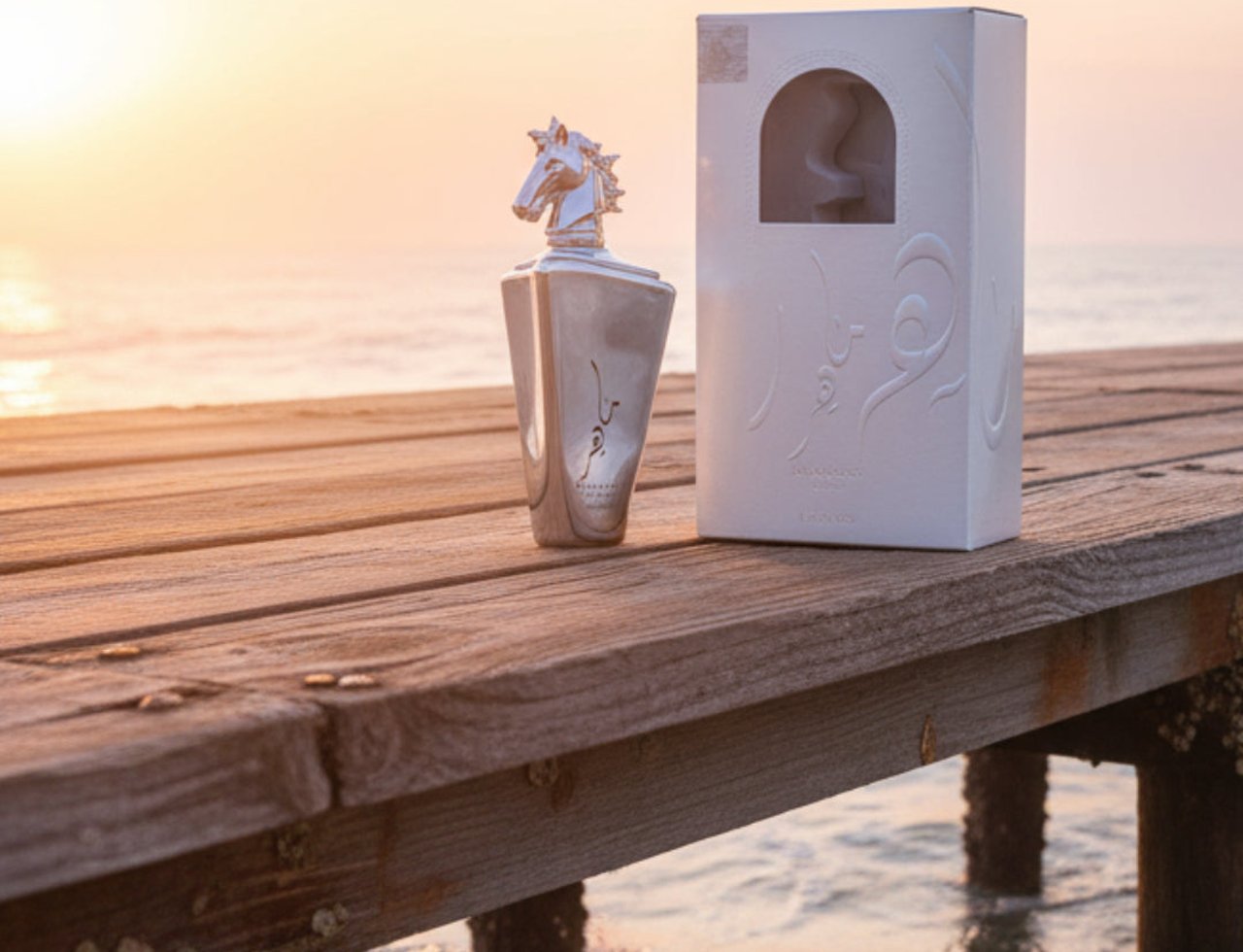 Lattafa Maahir Legacy by Lattafa 100ml EDP Man — Silver perfume bottle with horse head cap and white box on a wooden pier at sunset.