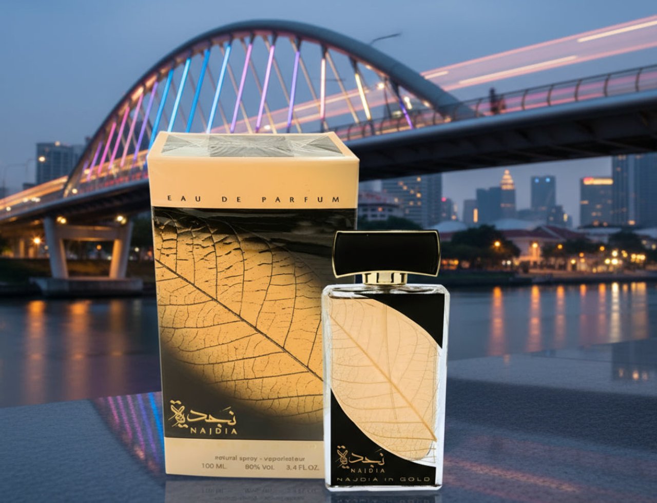 Lattafa Najdia in Gold by Lattafa 100ml Unisex — A perfume bottle and its box are displayed on a reflective surface with a blurred city bridge and skyline in the background.
