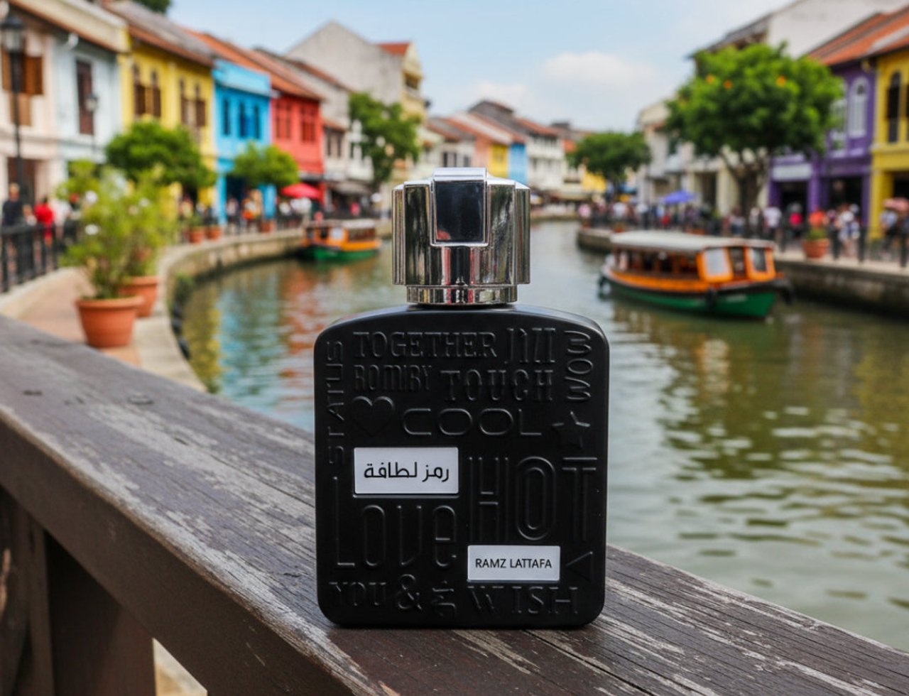 Lattafa Ramz Silver by Lattafa 100ml EDP for Men — Black perfume bottle with silver cap, embossed text, and Arabic script, placed on a wooden railing with a canal and colorful buildings in the background.