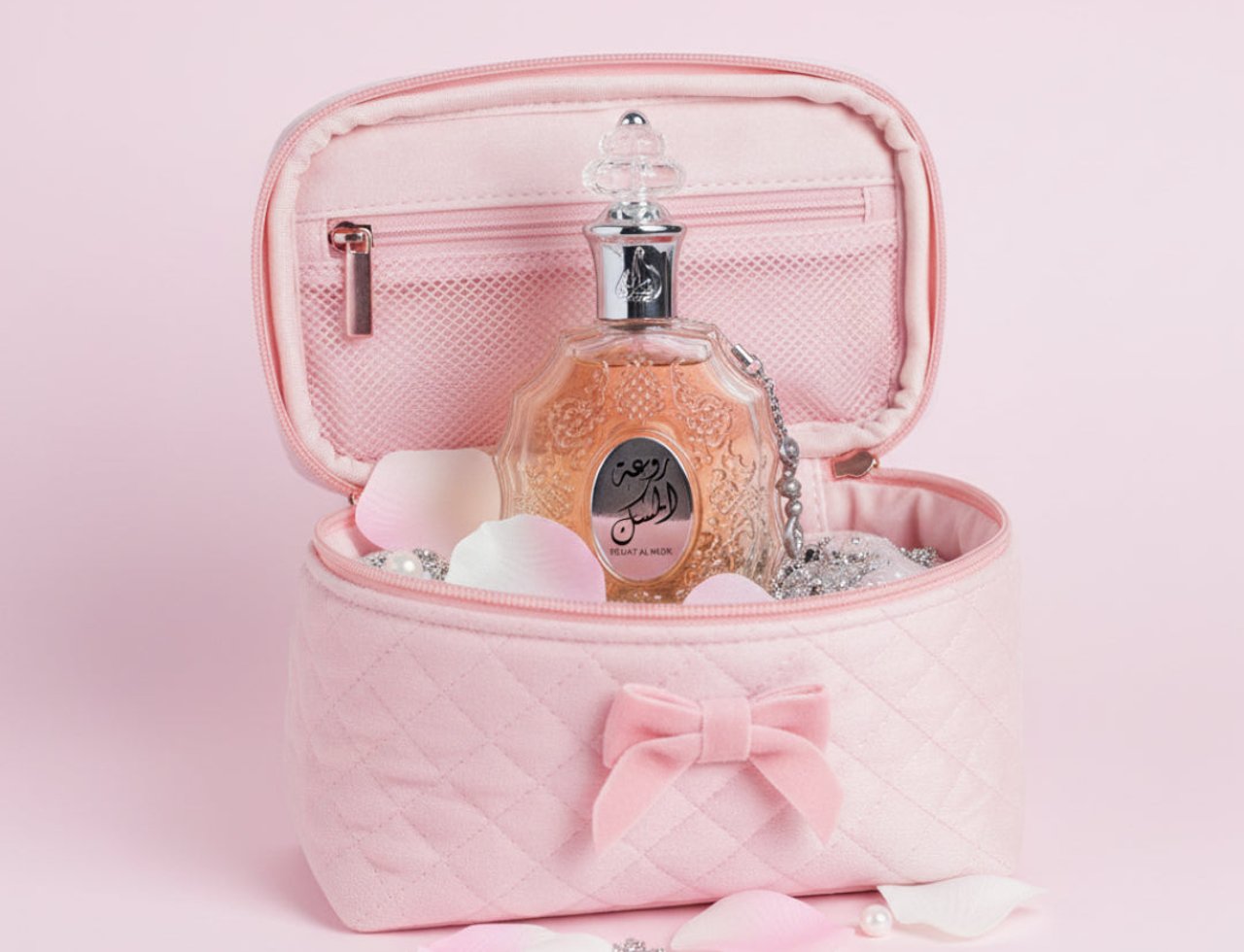 Lattafa Rouat Al Musk by Lattafa 100ml untuk Wanita — Beg kosmetik baldu merah jambu berkuil terbuka, memaparkan botol minyak wangi kaca jernih dengan perincian emas berukir dan rantai perak, dikelilingi kelopak mawar merah jambu dan mutiara di latar belakang merah jambu.