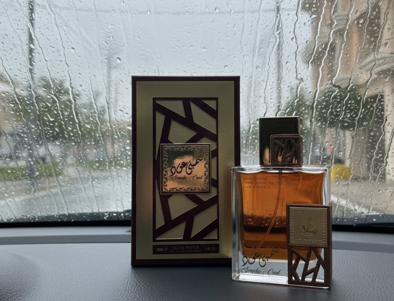 Lattafa Simply Oud 100ml EDP for Men — A glass perfume bottle with amber liquid and a decorative gold and brown cap, next to its matching rectangular box with a geometric design, displayed on a dark surface with raindrops on a window in the background.