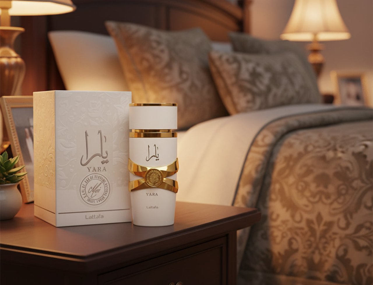 Lattafa Yara Moi by Lattafa 100ml EDP Women — Botol minyak wangi putih dengan perincian emas dan reben, di sebelah kotak putih berteksturnya, di atas meja sisi kayu gelap dengan katil dan lampu di latar belakang.
