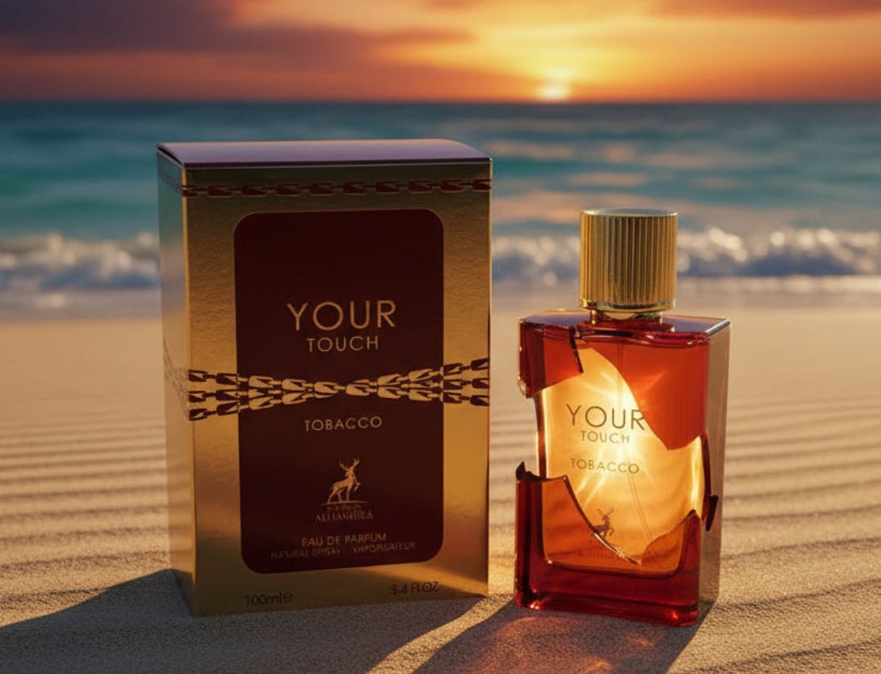 Maison Alhambra Tobacco Your Touch – Kotak emas dan botol minyak wangi kaca berpenutup emas terletak di pantai berpasir waktu matahari terbenam, dengan laut di latar belakang.