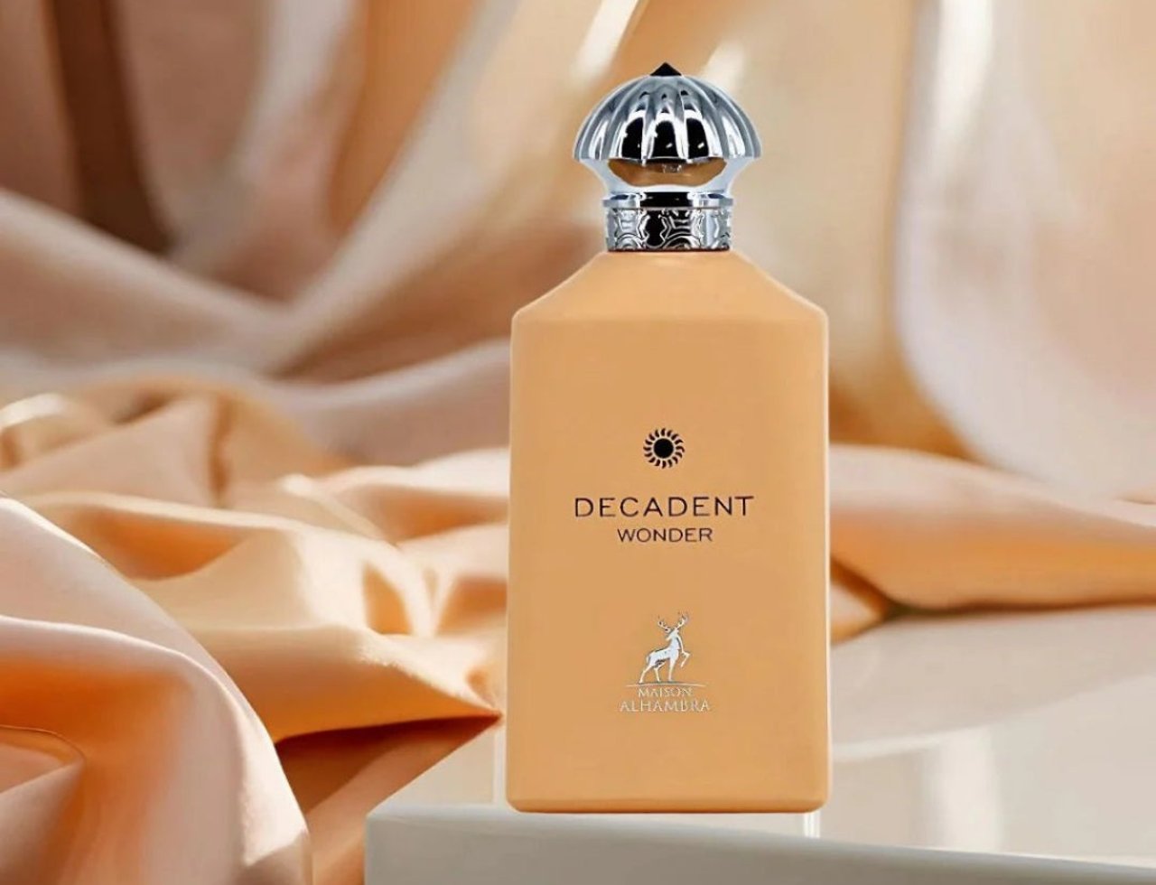 Maison Alhambra Decadent Wonder Her EDP 100 ml Alhambra — A peach-colored perfume bottle with a silver fluted cap, set against a backdrop of flowing peach satin fabric.
