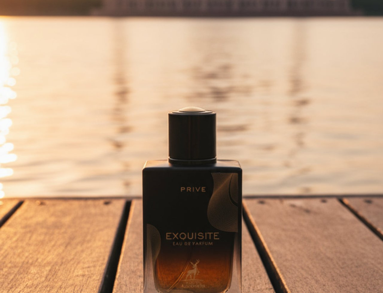 Maison Alhambra Exquisite Prive Maison Alhambra 100 ml EDP Men — A black and clear glass perfume bottle with gold accents sits on a wooden deck, with a blurred mosque and sunset over water in the background.