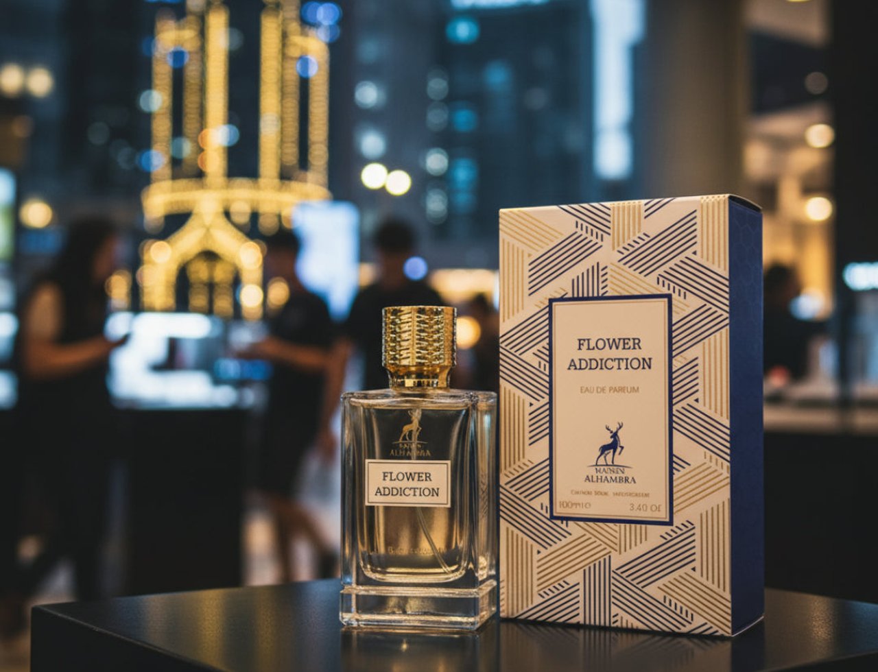 Maison Alhambra Flower Addiction by Maison Alhambra for Him 100ml — Clear glass perfume bottle with gold cap and white label, next to its blue and gold patterned box, with a blurred city lights background.