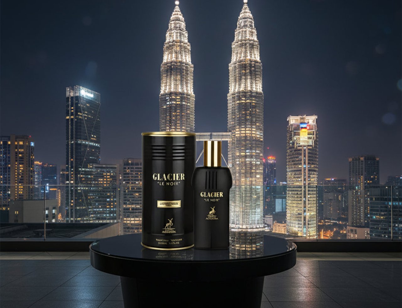 Maison Alhambra Glacier Le Noir — Botol minyak wangi hitam dan kotak silinder beraksen emas dipamerkan di atas meja bulat berlatar belakang pencakar langit yang diterangi cahaya pada waktu malam.