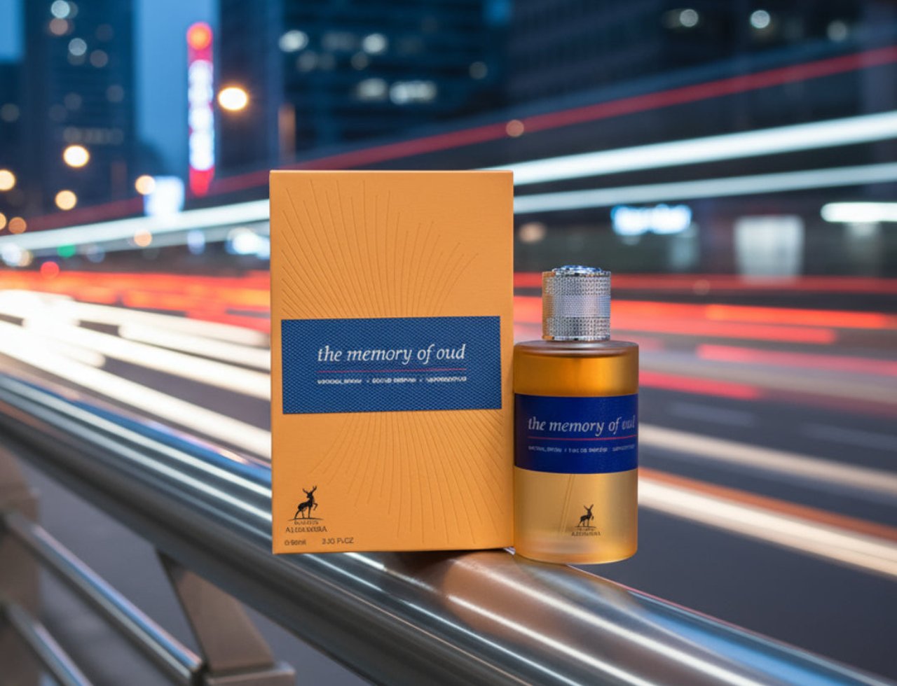 Maison Alhambra The Memory Of Oud EDP 100ml — Orange box and gold-capped perfume bottle displayed on a metal railing with a blurred city street at night in the background.