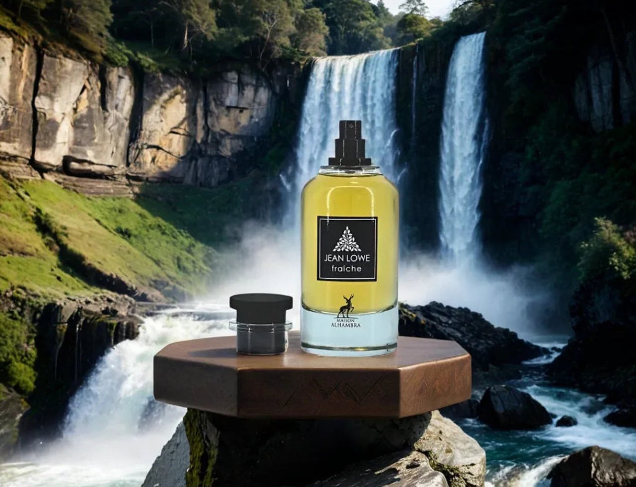 Maison Alhambra Jean Lowe Fraiche — Yellow perfume bottle and small black cap displayed on a wooden hexagonal stand in front of a majestic waterfall.