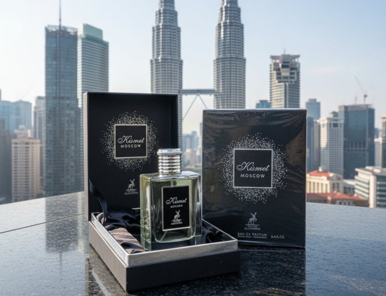 Maison Alhambra Kismet Moscow by Maison Alhambra 100ml EDP — Botol minyak wangi kaca jernih bertutup perak, dalam kotak hadiah hitam terbuka berlapik satin, bersebelahan kotak produk hitam yang sepadan, berlatar belakang pemandangan bandar dengan bangunan pencakar langit tinggi.